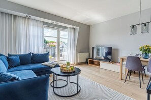 A modern living room with a blue sofa, a TV, a dining area with a table and chairs, and a view of buildings through large windows. - Imperial Apartments - Sopocki Parkur (Sopot)