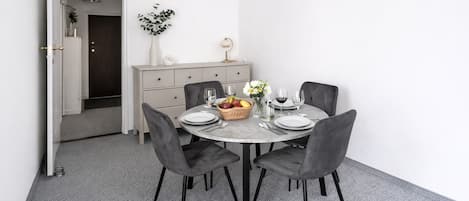 A dining area with a round table, four grey chairs, a white vase with flowers, a fruit basket, and wine glasses.