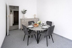 Ein Essbereich mit einem runden Tisch, vier grauen Stühlen, einer weißen Vase mit Blumen, einem Obstkorb und Weingläsern.