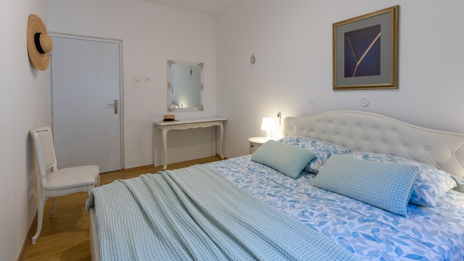 Ein Schlafzimmer mit Bett, Stuhl, Schreibtisch, Spiegel und einem gerahmten Bild an der Wand.