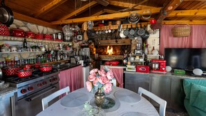 Een keuken met houten balken, een stenen muur en een open haard. Er staan rode pannen, een fornuis en een eettafel met stoelen.