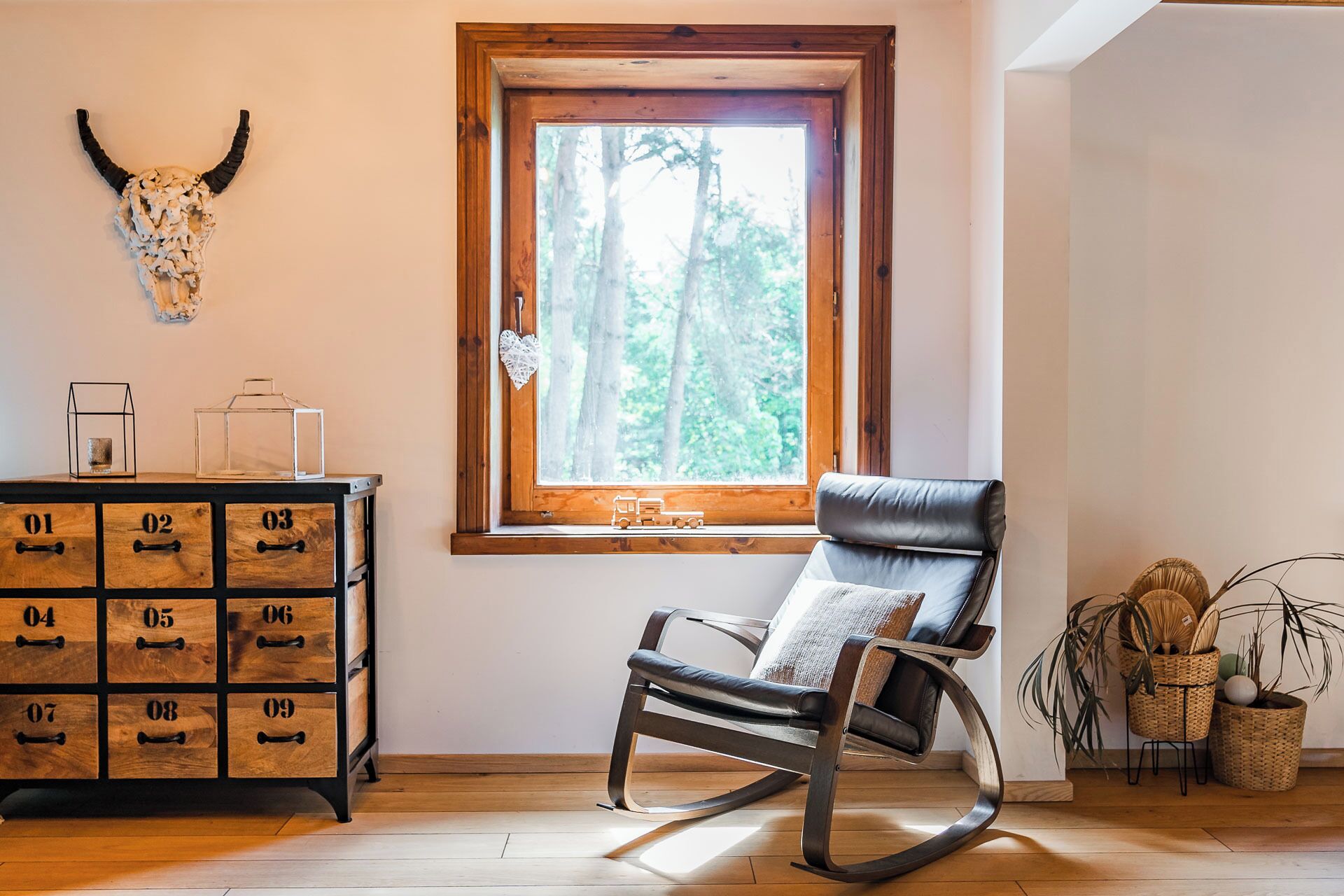 Una habitación con un aparador de madera, una mecedora, un ventanal con vistas a un bosque y un cráneo de toro colgado en la pared.