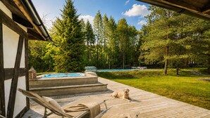 Terraza de madera con sillones de descanso y un jacuzzi, rodeada de árboles y una piscina.