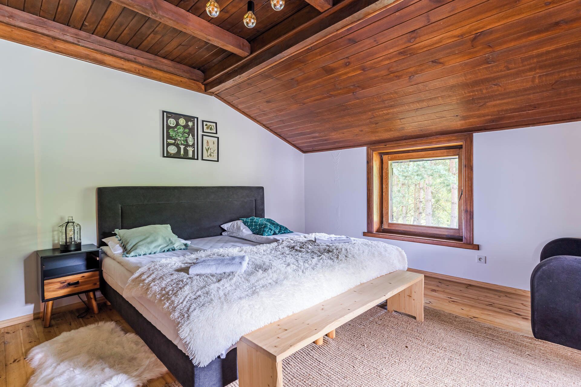 A bedroom with a wooden ceiling, a bed with a bench, a nightstand, a window, and wall art.