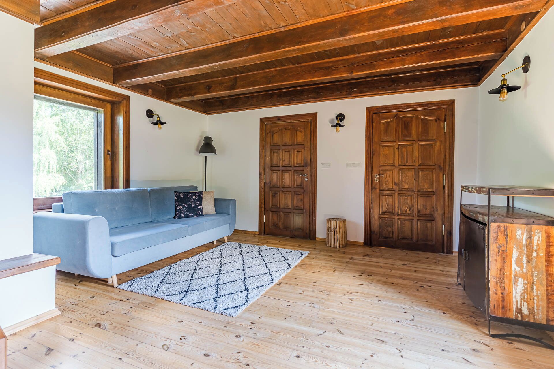 A room with a wooden ceiling, a blue sofa, a patterned rug, and two wooden doors.