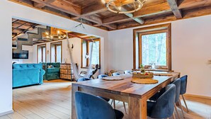 Un comedor espacioso con una mesa y sillas de madera, un gran ventanal con vistas a árboles, y una sala de estar acogedora con un sofá azul y vigas de madera en el techo.