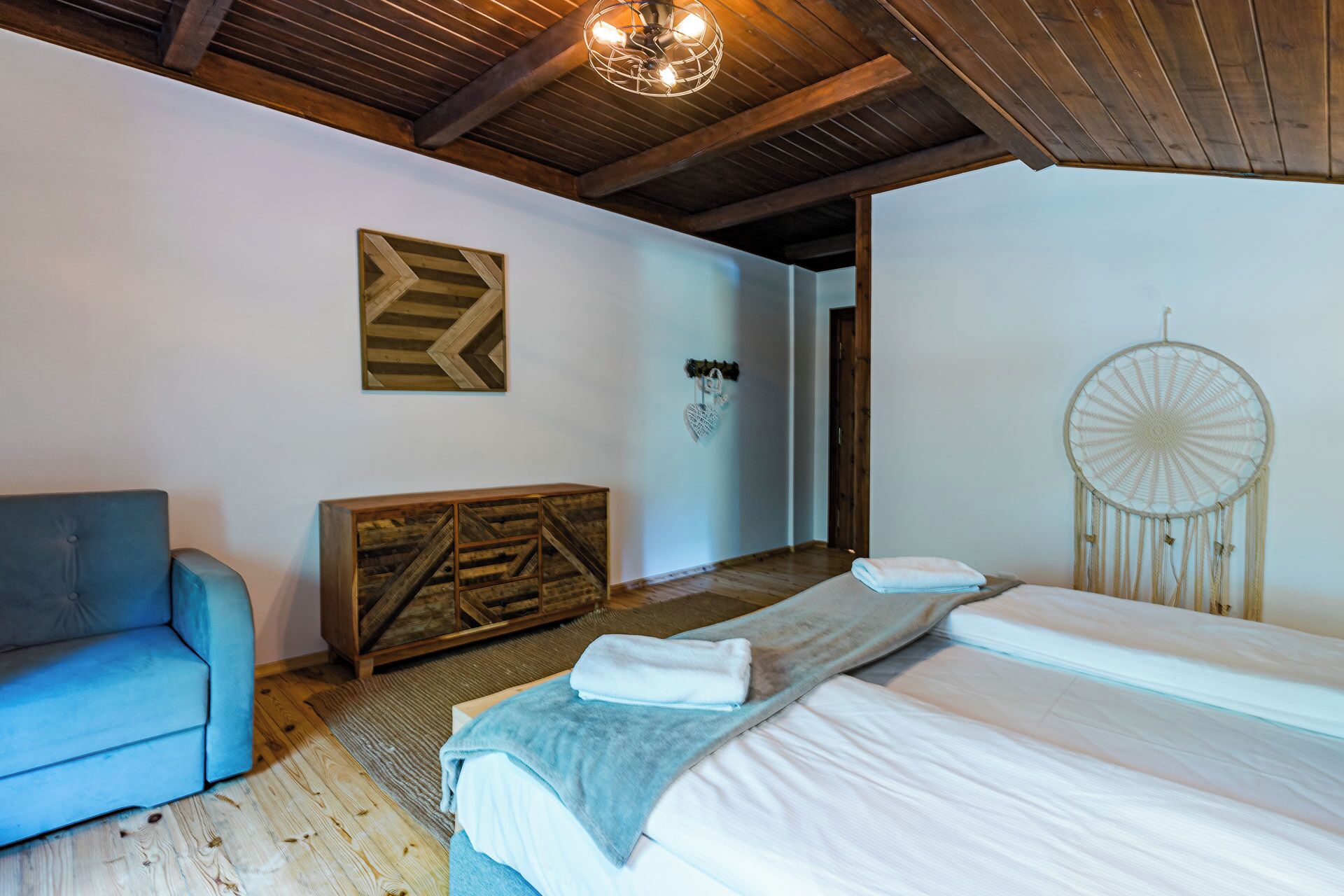 A bedroom with a bed, a blue chair, a wooden headboard, a dreamcatcher, and a wall with a geometric pattern.