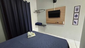 A hotel room with a bed, a TV mounted on a wooden panel, a towel on the bed, a shelf with a towel, and wall art.