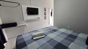 A bedroom with a checkered floor, a flat-screen TV mounted on the wall, a bed with a rolled-up white towel, and a door with a mirror.