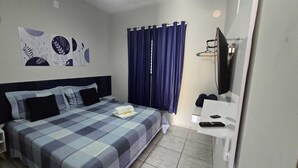 A hotel room with a bed, a desk with a TV, a window with curtains, and artwork on the wall.
