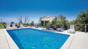 Ein rechteckiger Pool mit klarem, blauem Wasser, umgeben von Liegestühlen und Sonnenschirmen, vor einer Kulisse aus Bäumen und blauem Himmel.