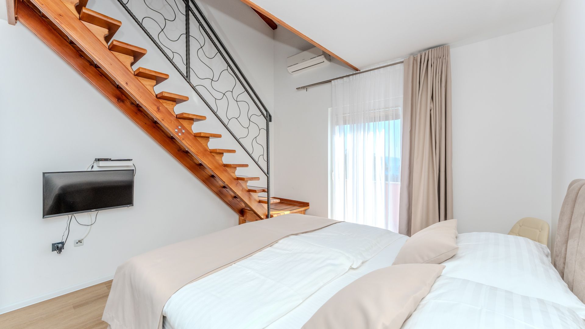 A bedroom with a wooden staircase, a bed with white bedding, a flat-screen TV mounted on the wall, and an air conditioning unit.