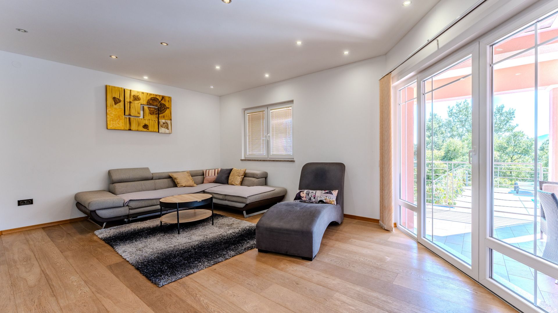 A modern living room with a sectional sofa, a grey armchair, and a coffee table. There is a large window providing a view of the outdoors.