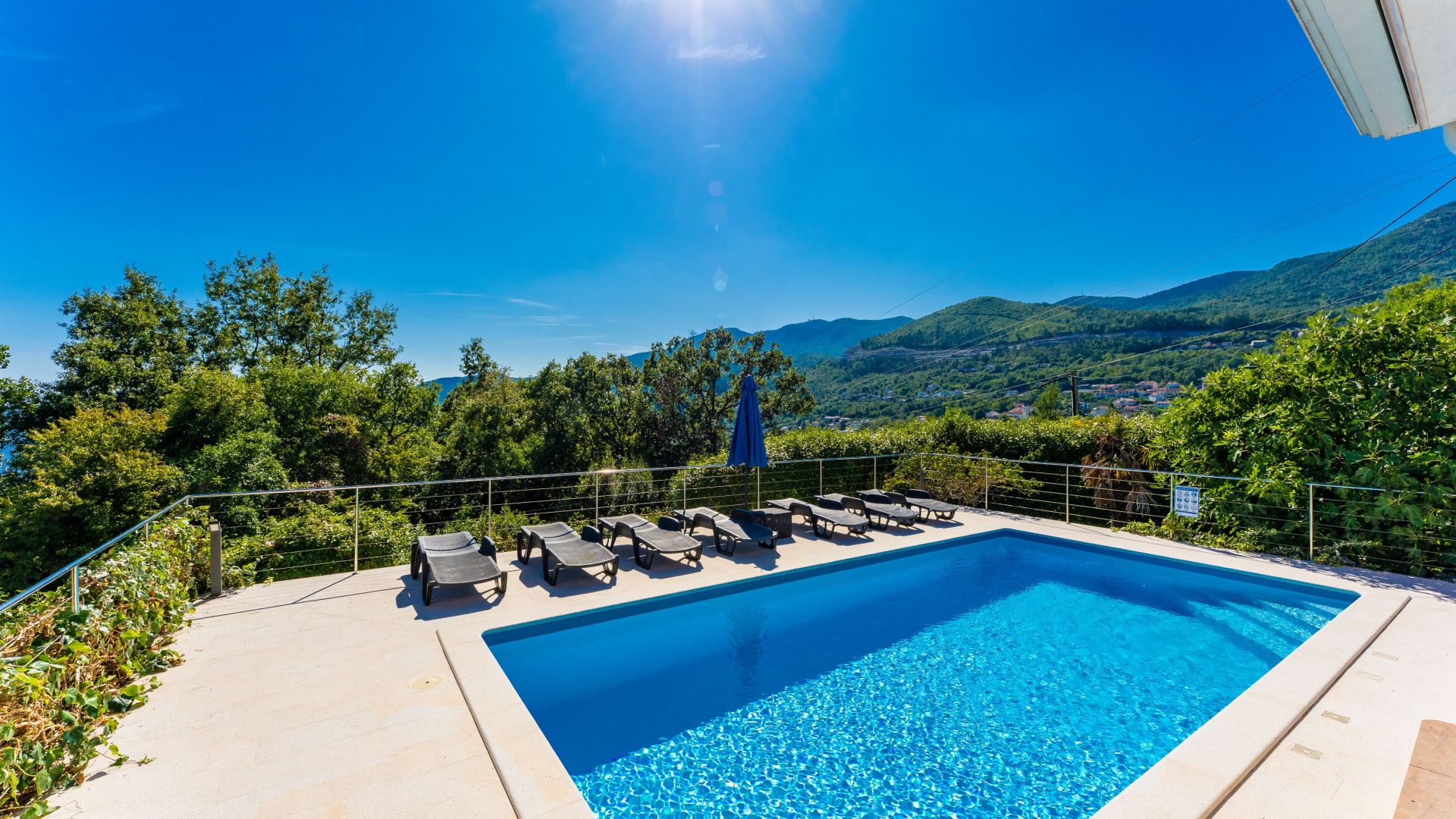 A rectangular swimming pool with clear blue water, surrounded by a metal railing and equipped with lounge chairs and a blue umbrella.