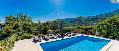 A rectangular swimming pool with clear blue water, surrounded by a metal railing and equipped with lounge chairs and a blue umbrella.