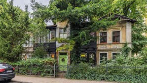 Un immeuble à plusieurs étages avec une porte verte, entouré d’une végétation luxuriante et d’une clôture.