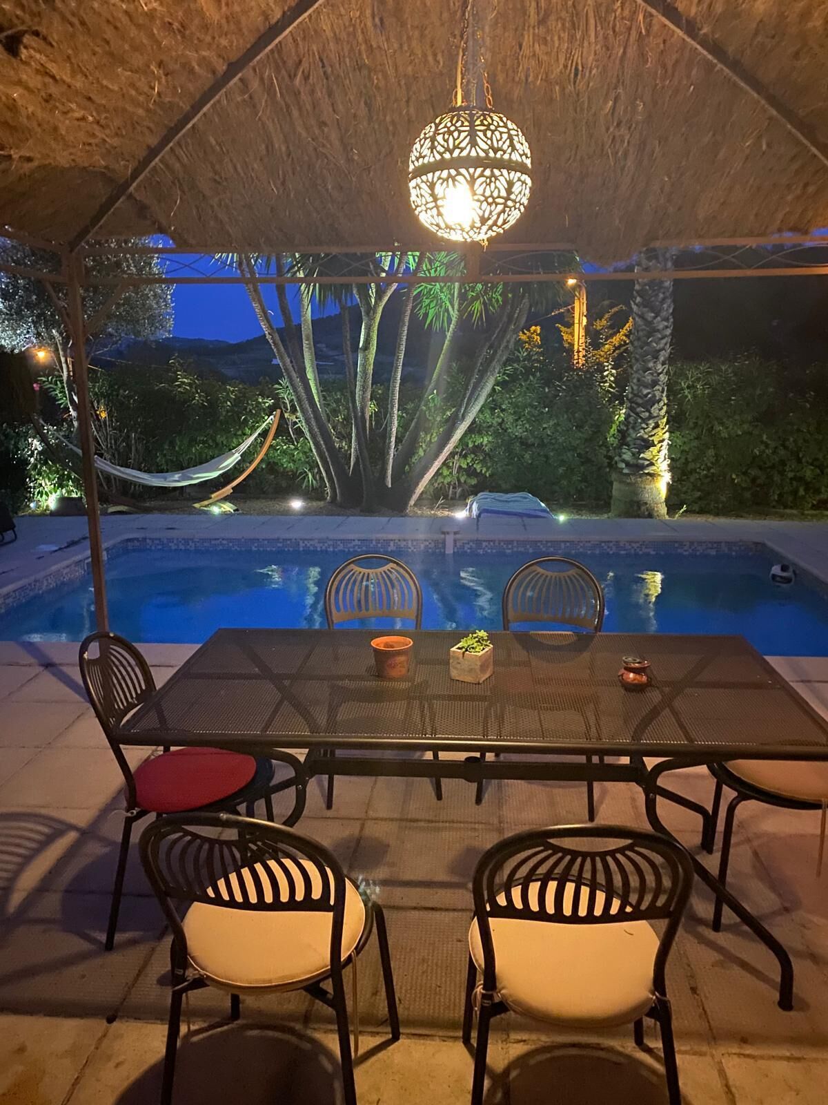 Area pranzo all'aperto con tavolo e sedie, un'amaca e vista su una piscina di notte.