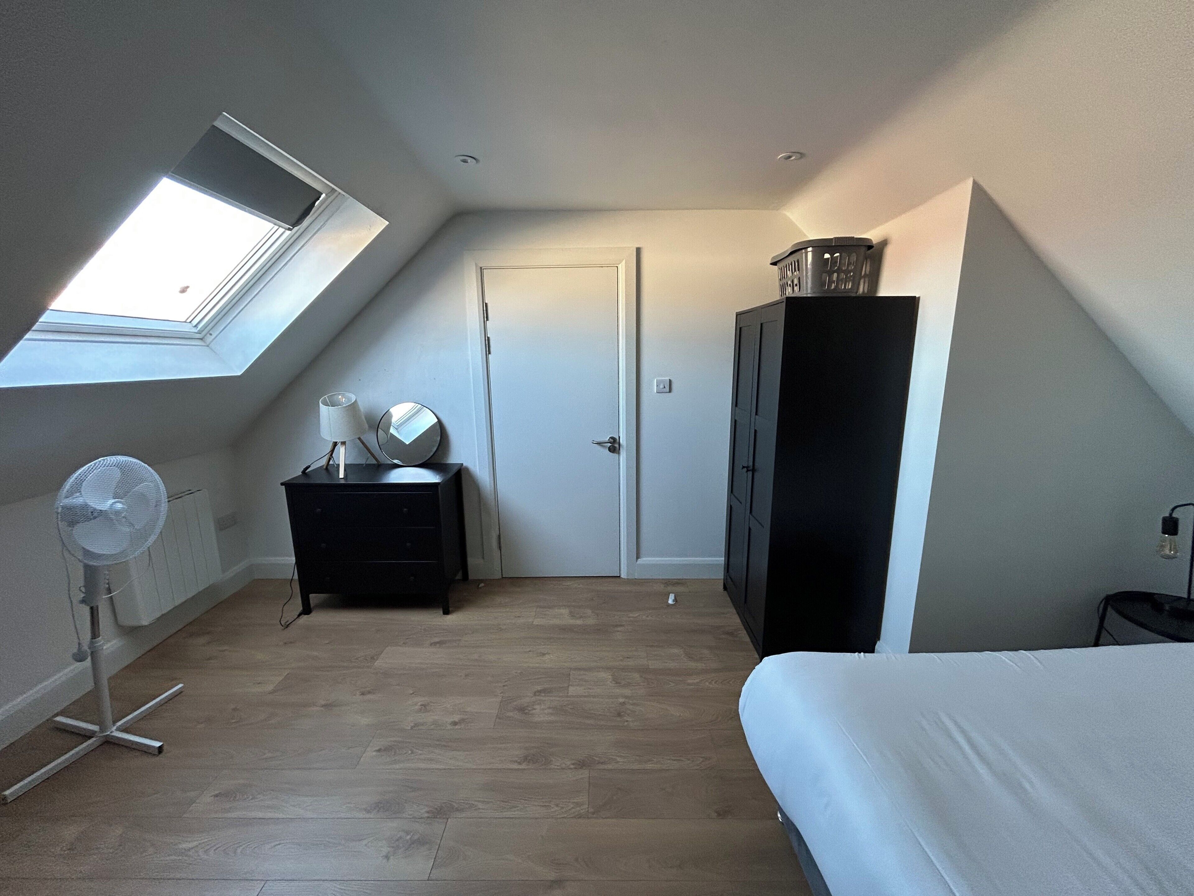 Ein Zimmer mit Oberlicht, einem Bett, einem Schminktisch, einem Spiegel, einem Ventilator und einem Kleiderschrank.