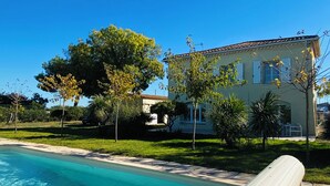 A swimming pool with clear blue water, a white poolside lounge, and a building with a tiled roof in the background. - JF IMMO-VILLA RARE GARDEN PRIVATE POOL 10 PEOPLE SEA VIEW V0861 (Agde)