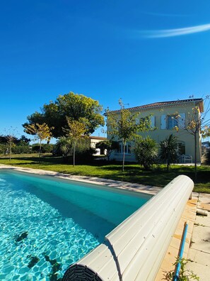 Une piscine à l’eau d’un bleu cristallin, un salon de jardin blanc situé au bord de la piscine et, au fond, un bâtiment dont le toit est recouvert de tuiles.