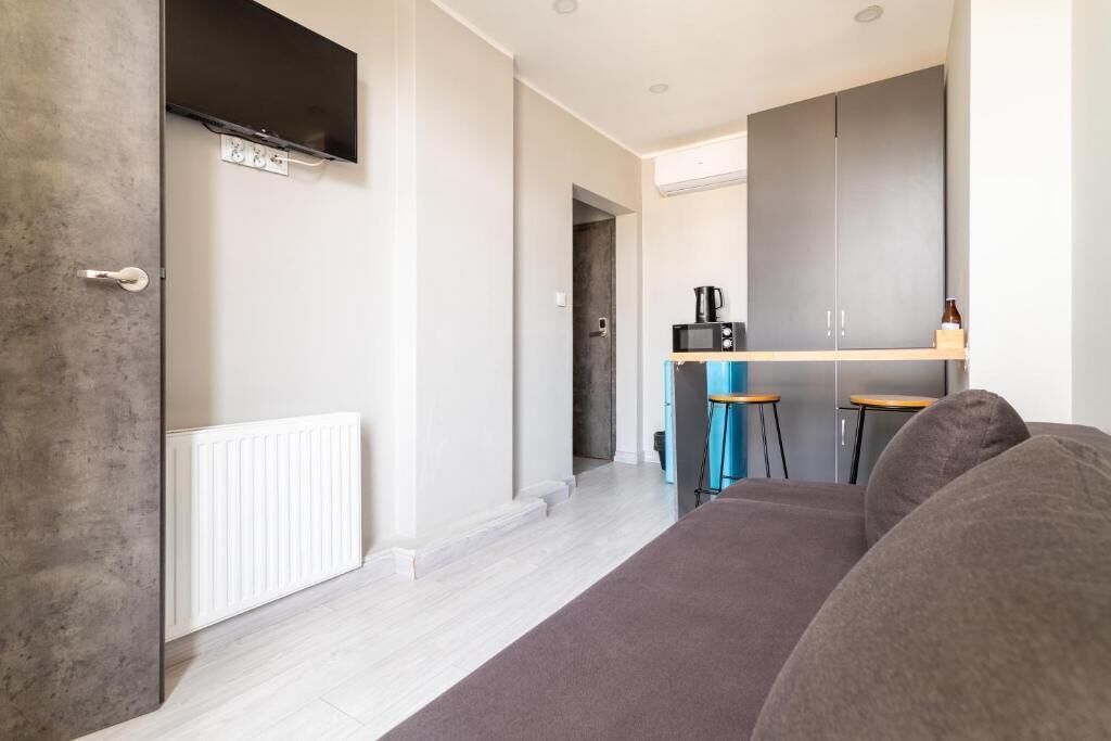 A modern living room with a sofa, a television mounted on the wall, a kitchen area with a microwave, and a radiator.