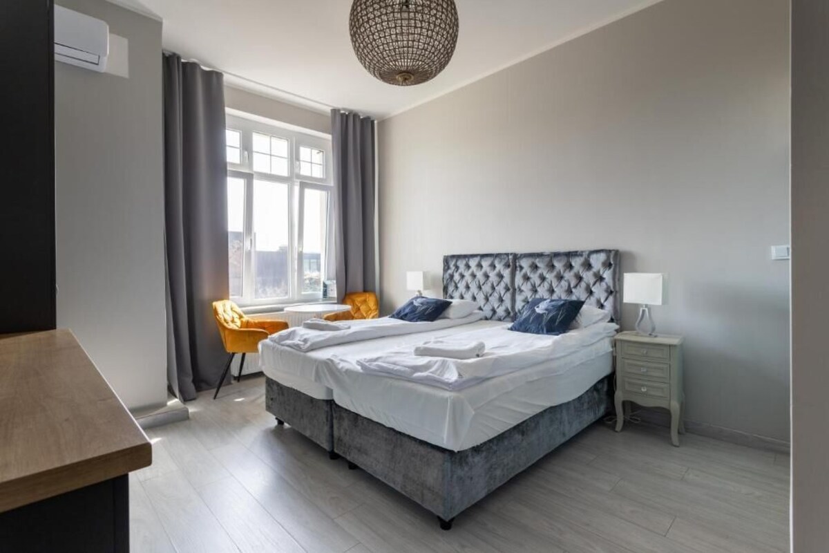A double bed with a tufted headboard, a bedside table with a lamp, a chair, and a window with curtains.