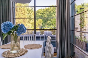 A dining area with a table, chairs, a vase with flowers, and a view of a balcony with a sofa and railing. - Imperial Apartments - Bernadowska (Gdynia)