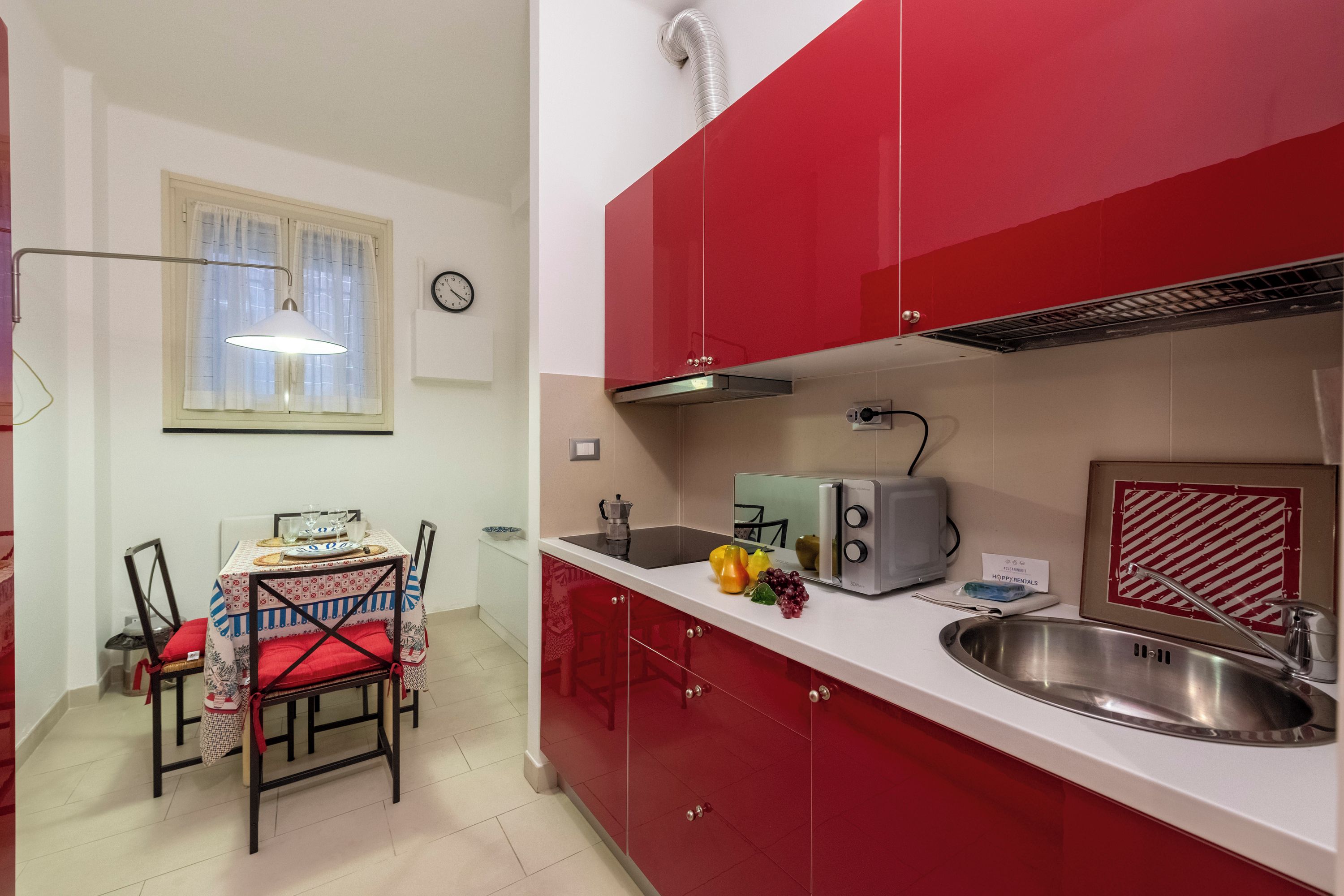 A compact kitchen with red cabinets, a white countertop, a stainless steel sink, a toaster, and a window with curtains.