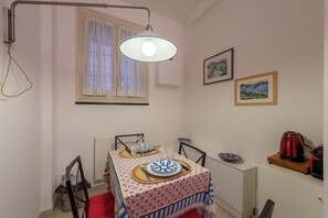 Ein Essbereich mit Tisch für zwei Personen, einem fensterbelaubten Fenster, einer Wandlampe und einem roten Wasserkocher auf einem Holftablett.