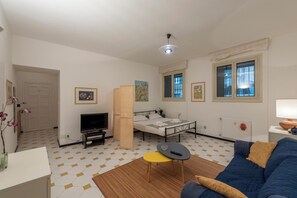 Ein Zimmer mit einem Bett, einem Fernseher, einem Sofa, einem kleinen Tisch und einem Fenster mit Jalousien.
