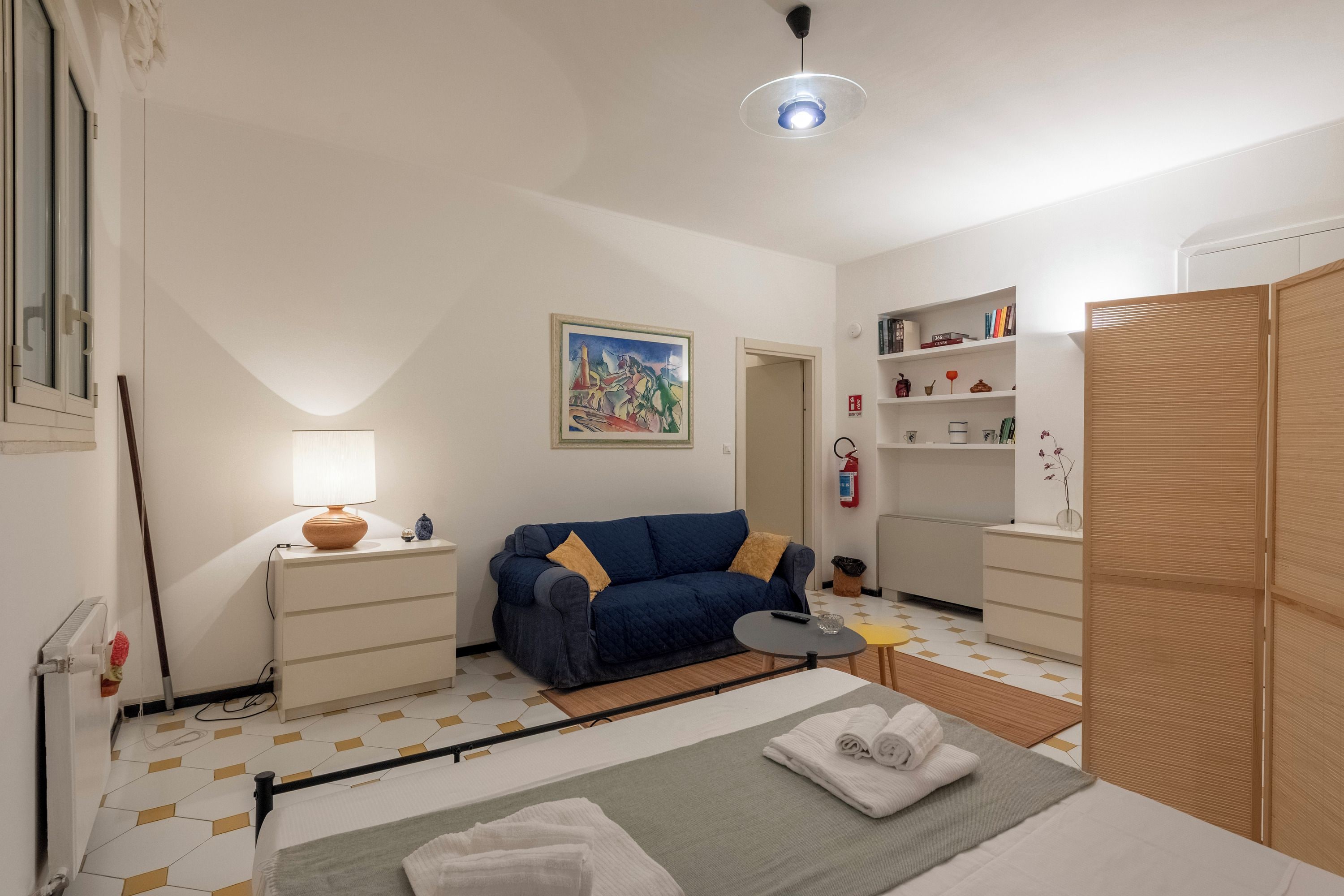 Una stanza con un letto, un divano, un tavolino, una lampada, un quadro, una libreria e un armadio.