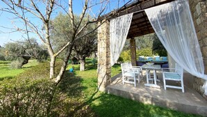 Un área de estar al aire libre con techo, mobiliario blanco, un edificio de piedra y un árbol sin hojas.
