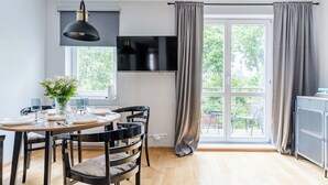 Un coin repas avec une table dressée pour quatre personnes, un téléviseur fixé au mur au-dessus, et une grande fenêtre avec des rideaux.