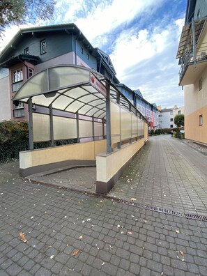 Ein überdachter Gang mit einem Schild, einem gepflasterten Weg und Gebäuden im Hintergrund.