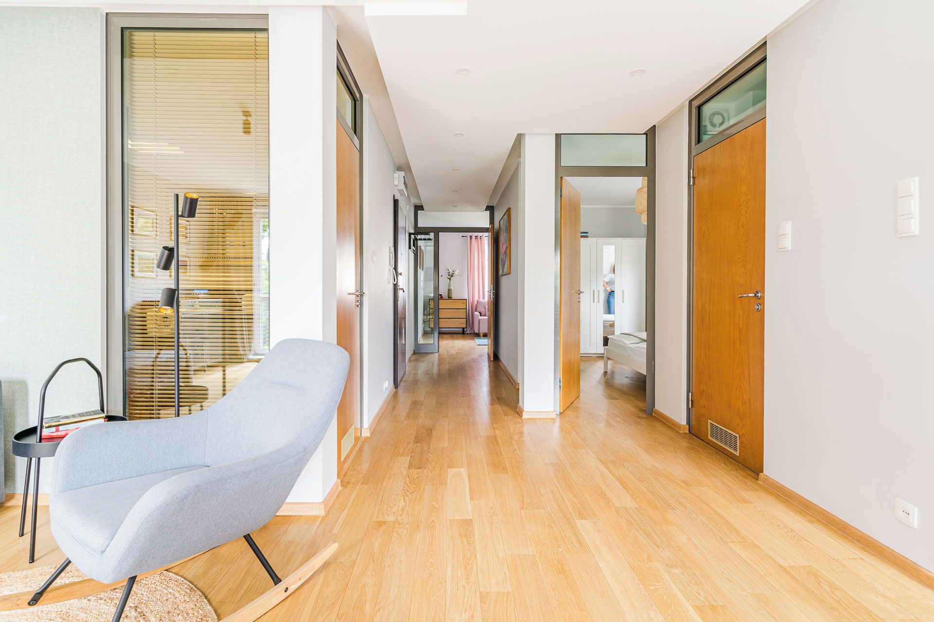A modern interior with a wooden floor, a grey lounge chair, and a glass door leading to a bathroom.