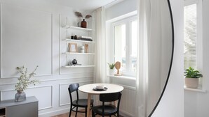 Ein Zimmer mit einem runden Tisch, zwei Stühlen, einem Bücherregal, einem Fenster mit Vorhängen und einer Topfpflanze.