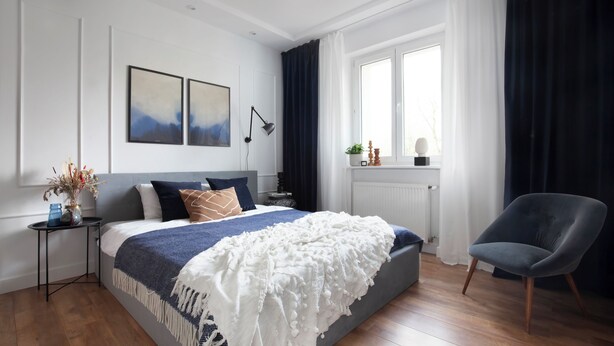 Ein Schlafzimmer mit einem großen Bett, einem Nachttisch, einem Sessel, einem Fenster mit Vorhängen und Bildern an der Wand.