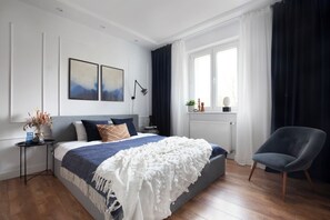 Un dormitorio con una cama grande, una mesita de noche, una silla, un ventanal con cortinas y una obra de arte en la pared.