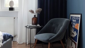 Ein Zimmer mit Fenster, einem Sessel, einem kleinen Tisch mit Vase und Büchern sowie einem Bild an der Wand.