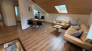 En rummelig loftrum med træbjælker, et spiseområde med et træbord og stole, en sofa og et lille bord med en lampe.