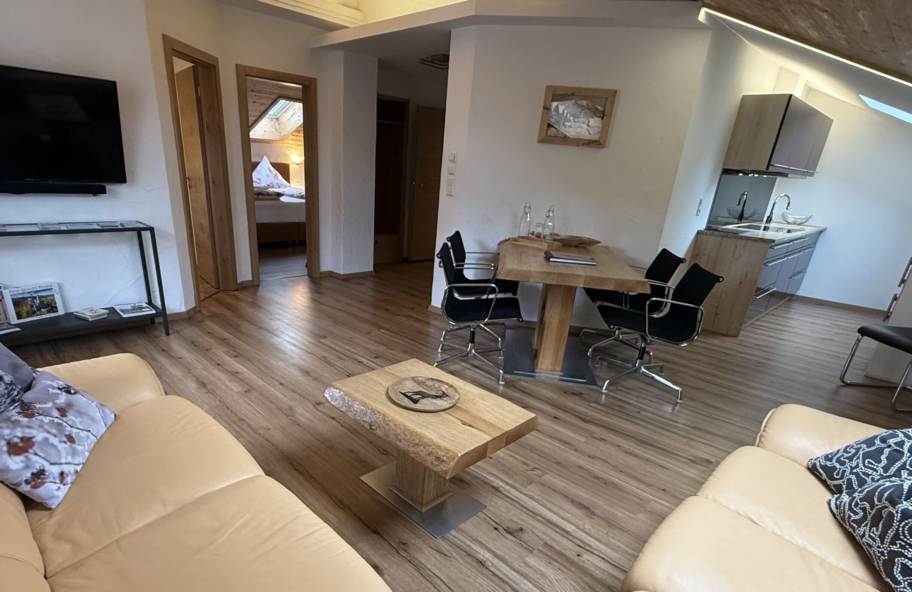 Een moderne woonkamer met een houten salontafel, beige banken, een eethoek met een houten tafel en stoelen, een keuken met een fornuis en kastjes, en een tv die aan de muur is bevestigd.