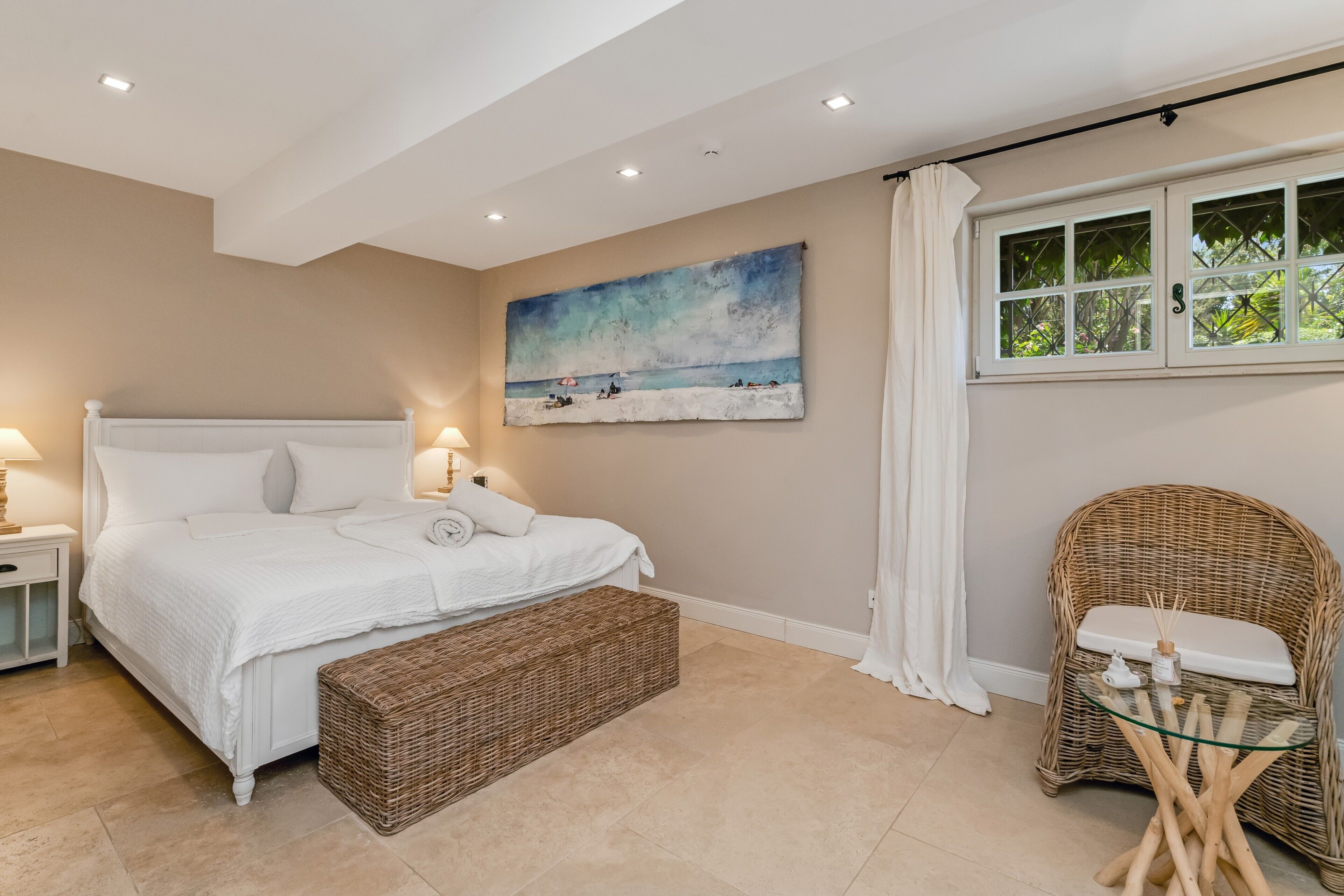 Ein Schlafzimmer mit einem Bett, einer Korbsitzbank, einem Korbstuhl, einem Strandbild und einem Fenster mit Vorhängen.