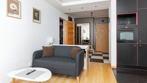 A modern living room with a grey sofa, a small white table, and a kitchen area with black cabinets and a wooden door. - RentPlanet - Apartament Skoroszewska (Warsaw)