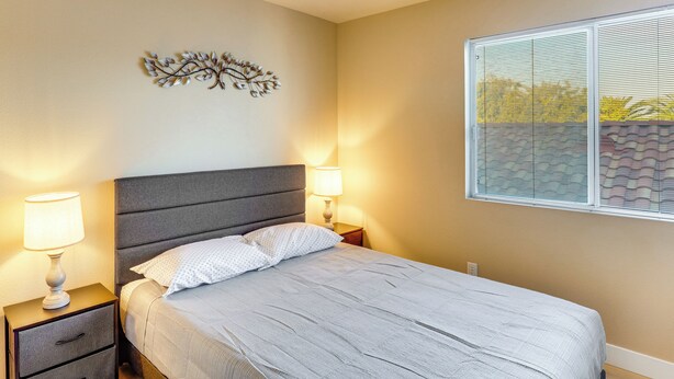 Un dormitorio con cama, dos mesitas de noche con lámparas, un adorno en la pared y una ventana con persianas.