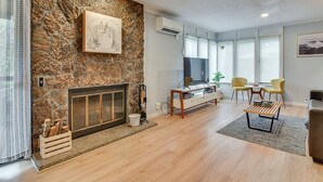 A modern living room with a stone fireplace, a flat-screen TV, wooden flooring, and a dining area with yellow chairs. - Relax Poolside! All-season Base in Mt Hood Village (Welches)