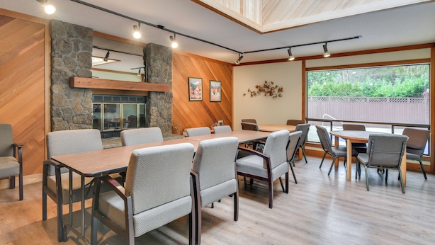 A conference room with a wooden table, grey upholstered chairs, a stone accent wall, and a large window with a view of greenery. - Relax Poolside! All-season Base in Mt Hood Village (Welches)
