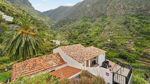A white building with a tiled roof, a balcony, and a view of a mountainous valley with terraced fields.