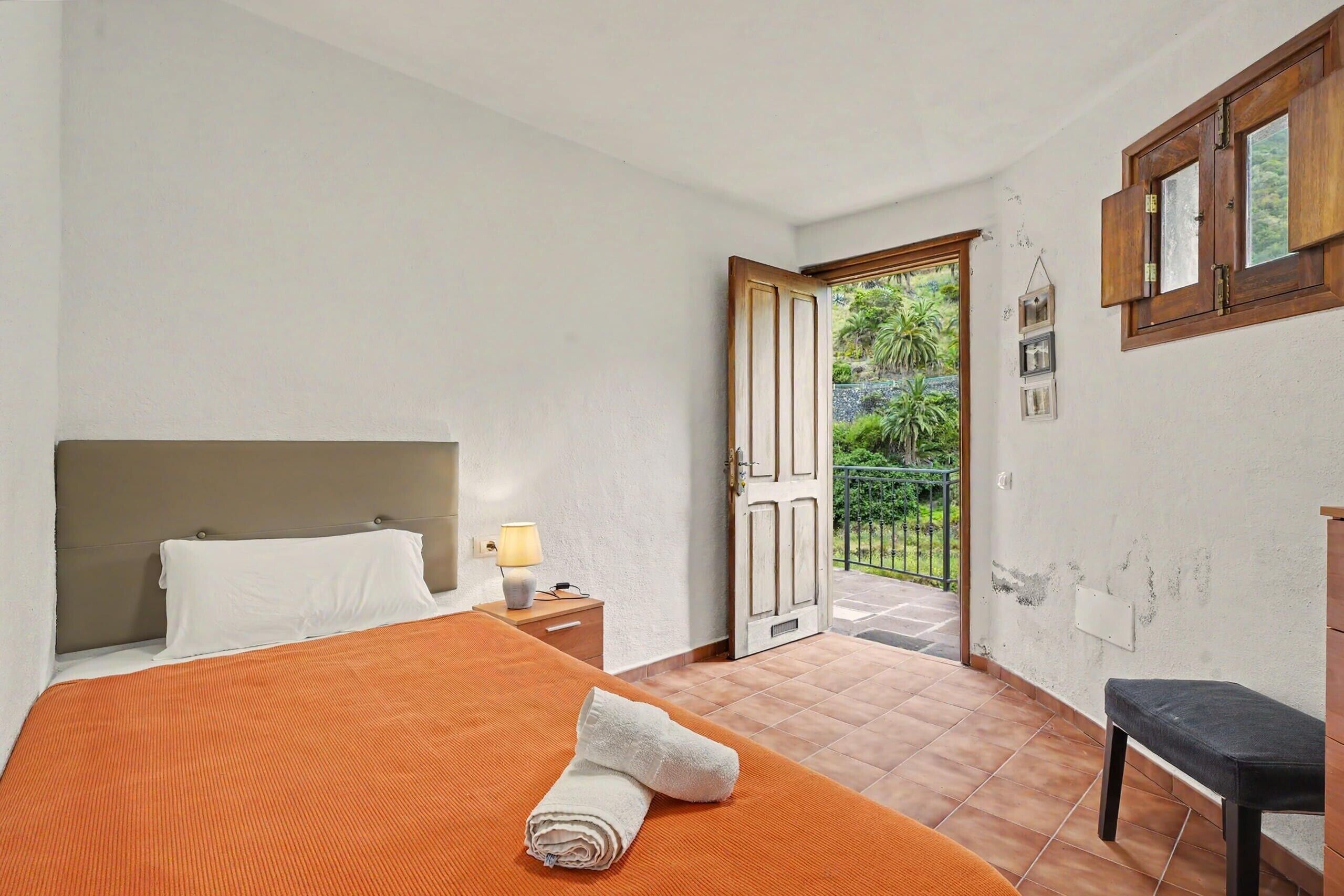 Une chambre d’hôtel avec un lit, une table de chevet, une lampe, un banc, une porte donnant sur un balcon et une vue sur la verdure environnante.