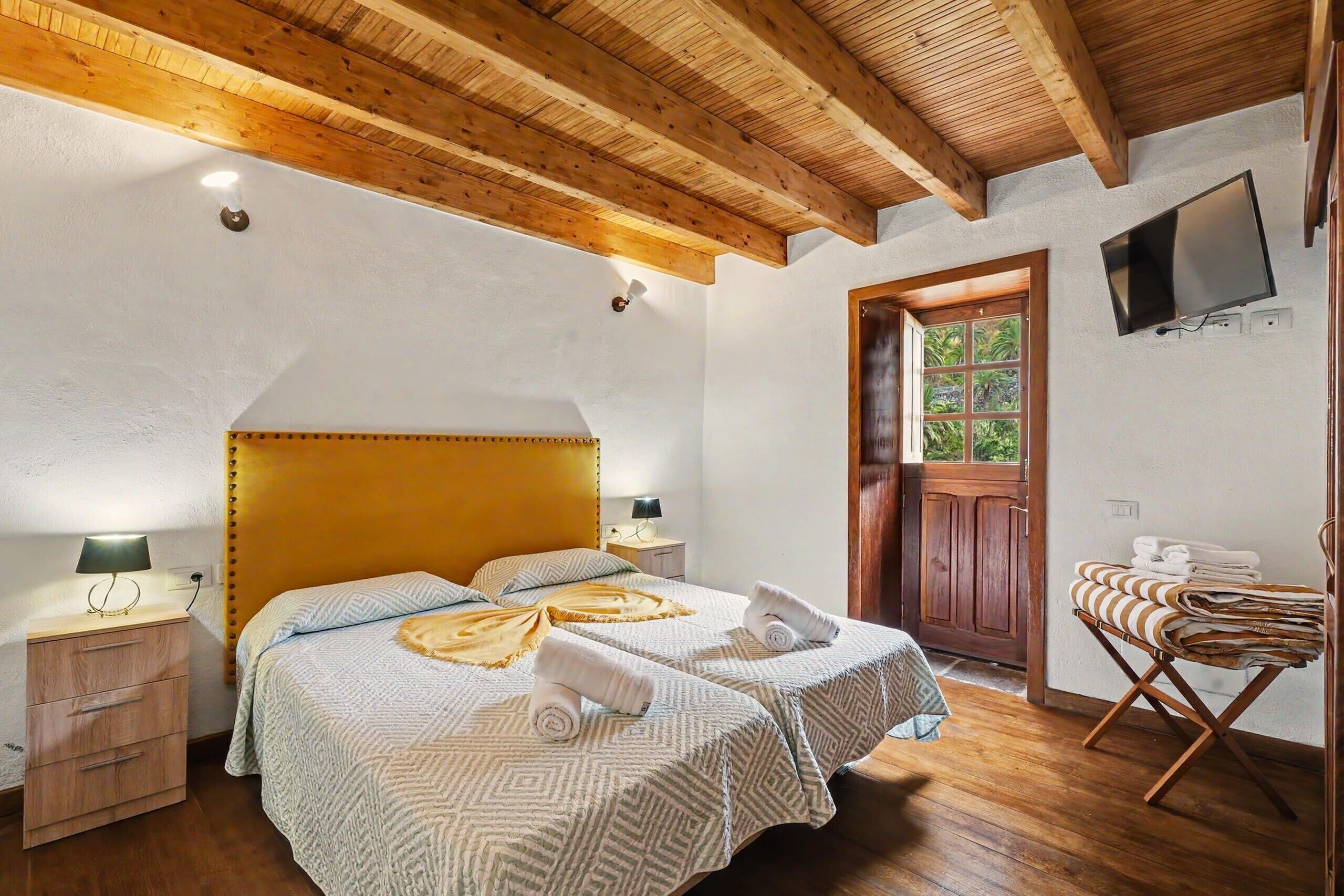 Een slaapkamer met houten plafond, een bed met beddengoed met patroon, een nachtkastje met lamp en een aan de muur bevestigde televisie.