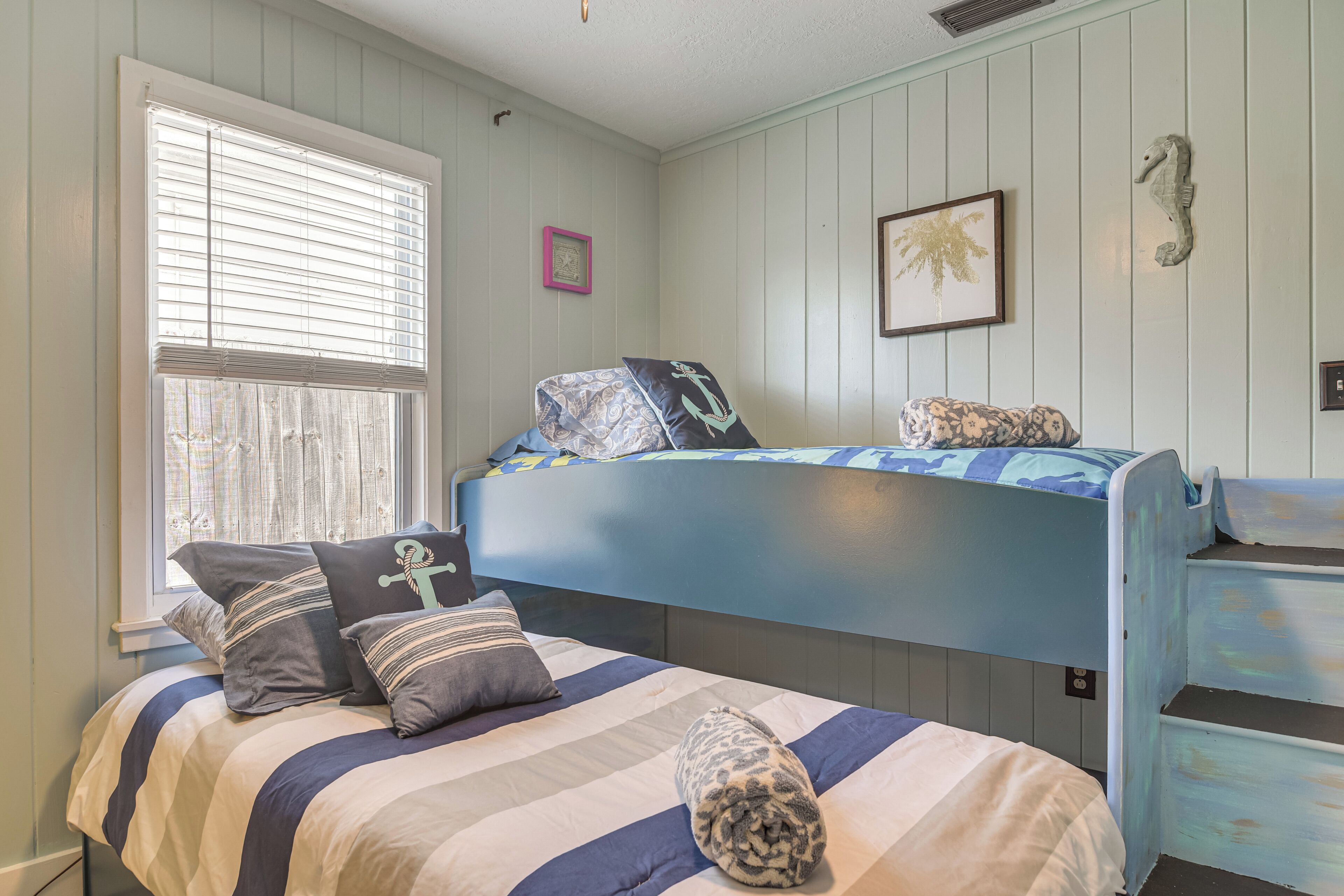 Un dormitorio con una cama nido, una ventana con persianas, un cuadro enmarcado y un adorno con forma de caballito de mar en la pared.